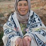 Une femme en tenue paysanne avec une veste berbère en laine sourie et montre le bout de ses doigts de couleur bleue