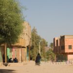 La silhouette d'une femme Aït Atta enveloppée dans son voile noir brodé de laine vive dans une rue en terre bordée de maisons en terre crue et d'acacias